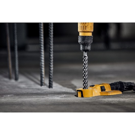 A yellow DEWALT drill with a spiral drill bit is being used to drill into a concrete surface. Metal rods are visible in the background, indicating a construction environment.