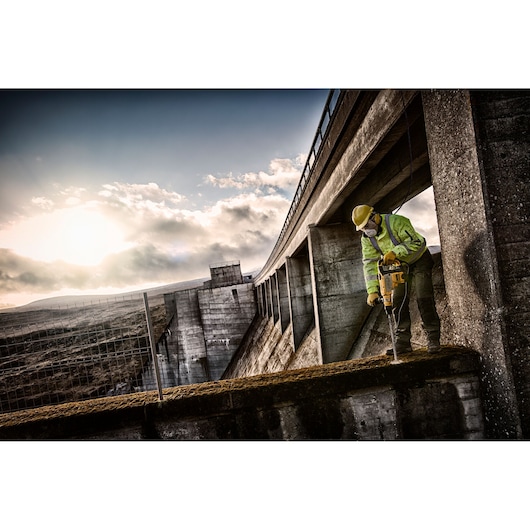 Worker using a DEWALT drill on a concrete structure outdoors, wearing safety gear under a cloudy sky.