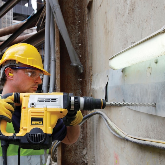 Worker uses DEWALT rotary hammer drill DT60829 to drill into a concrete wall on a construction site.