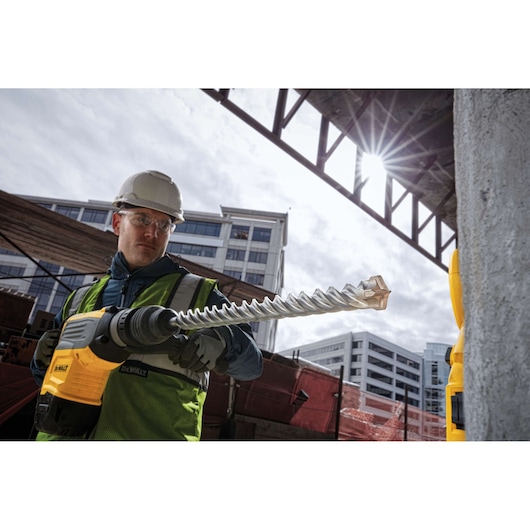 Worker using a DEWALT drill with a large bit at a construction site, wearing safety gear, with buildings in background.