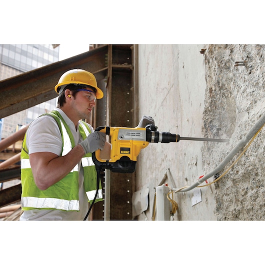 Worker uses DEWALT DT6825 chisel tool to break concrete wall at construction site, wearing safety gear.