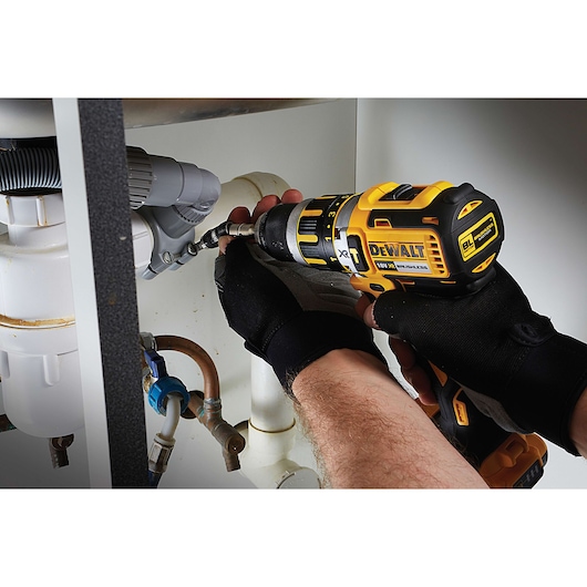 Person uses DEWALT drill to work on pipes under a sink, wearing black gloves for safety.