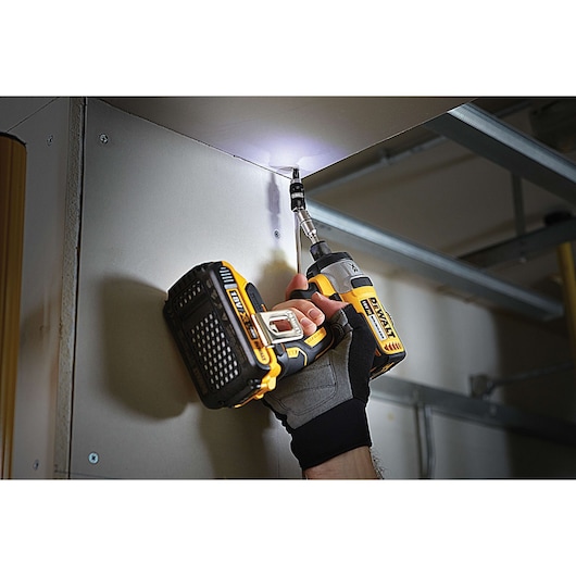 Hand uses a DEWALT cordless drill to secure a screw in a ceiling corner, tool emits light for visibility.