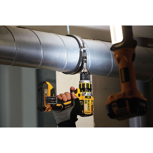 Hand uses a DEWALT cordless drill to fasten a screw into a metal pipe clamp in an industrial setting.