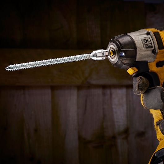 DEWALT power drill with a long screw attached, held by a gloved hand, used for woodworking or construction.
