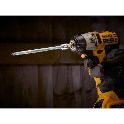 A DEWALT cordless drill with a long screw bit attached, held in a gloved hand, shown indoors.