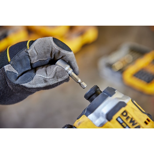 Gloved hand inserts a DEWALT bit into a yellow power tool. Tool kits are blurred in the background.