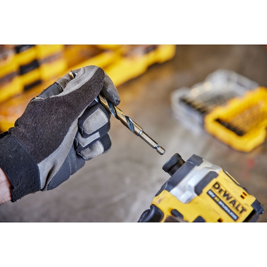 Gloved hand holding drill bit near DEWALT tool with yellow storage case in background.