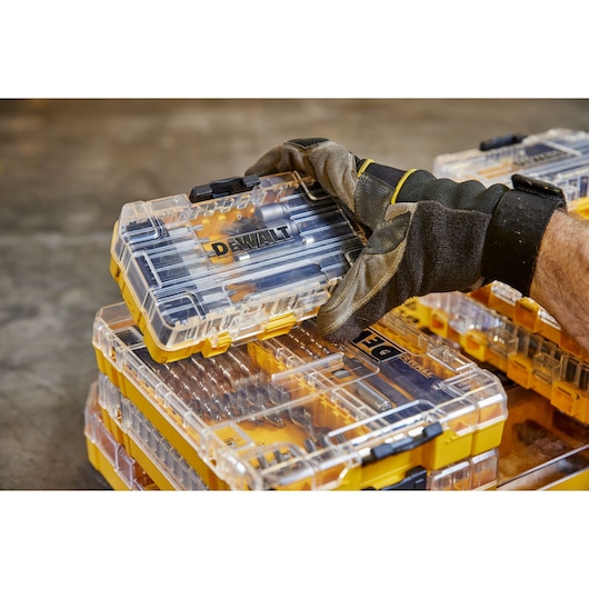 Gloved hand holding a DEWALT transparent tool case with drill bits and accessories stacked on a work surface.