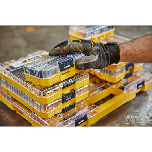 Gloved hand stacking yellow DEWALT storage cases with clear lids on a workshop floor.