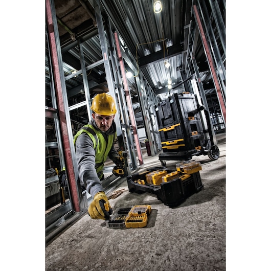 Worker in safety gear using DEWALT tools and bit set DT70716 at a construction site with toolboxes on the floor.