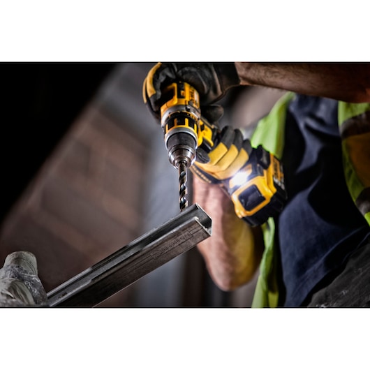 Close-up of a DEWALT drill being used to drill into a metal bar by a worker wearing gloves.