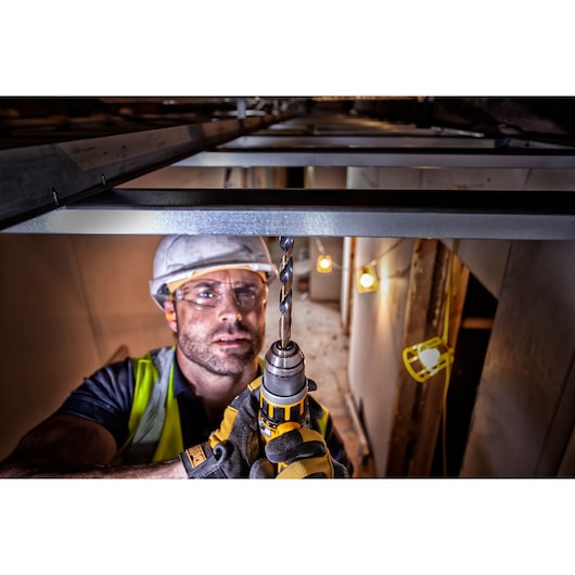 Worker uses DEWALT power drill to drill metal beam indoors, wearing safety gear and gloves.