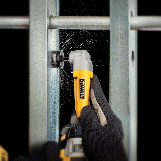 Gloved hand uses DEWALT right angle attachment to drill a hole in metal framing, producing metal shavings.