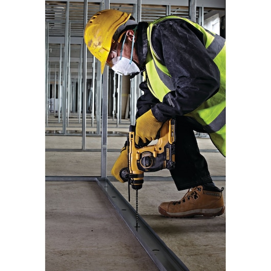 Worker uses a DEWALT drill on a metal frame, wearing safety gear and a mask at a construction site.