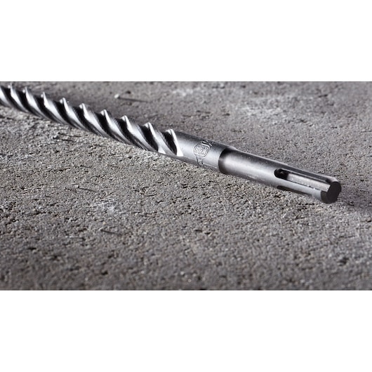 A close-up image of a DEWALT SDS-Plus masonry drill bit lying on a rough concrete surface.
