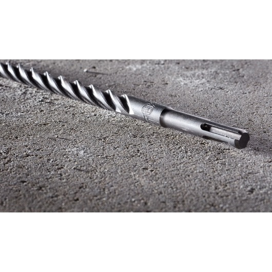 A close-up image of a DEWALT SDS drill bit with a spiral design, lying on a rough concrete surface.