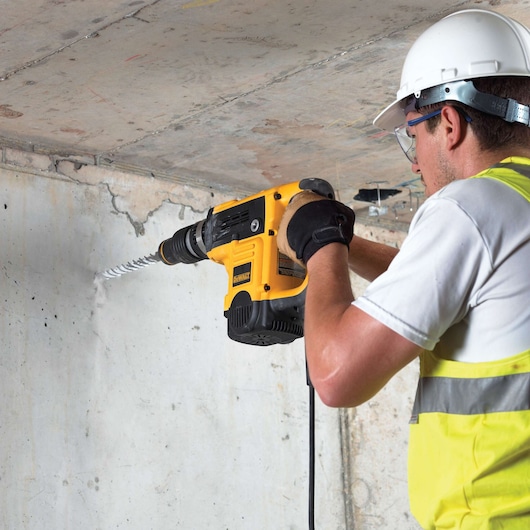 Worker uses a DEWALT hammer drill to drill into a concrete wall, wearing safety gear and high-visibility vest.