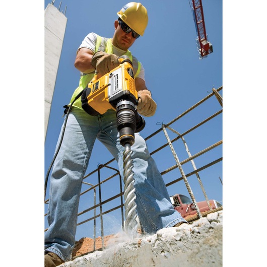 Worker uses a yellow DEWALT drill to bore into concrete at a construction site with rebar and a crane visible.