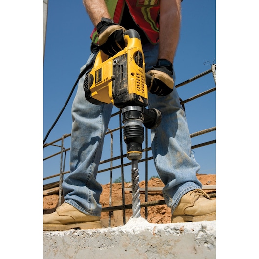 Worker uses a yellow DEWALT drill to bore into concrete at a construction site, wearing gloves and boots.