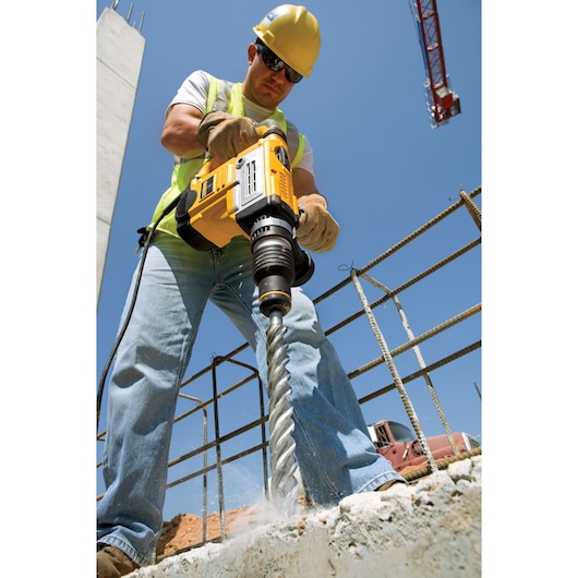 Worker using a DEWALT rotary hammer drill on concrete at a construction site under a clear blue sky.