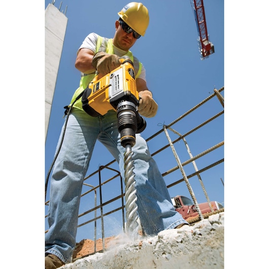 Person using a DEWALT rotary hammer drill on concrete at a construction site under clear blue sky.