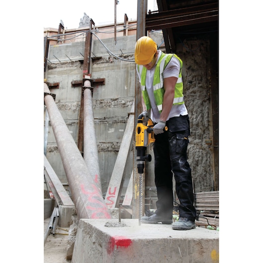 Worker uses DEWALT hammer drill DT9420_A7 on concrete at a construction site, wearing safety gear and helmet.