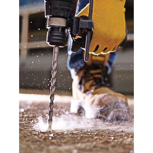 Close-up of a gloved hand using a drill on a concrete surface, with dust and safety boots visible.