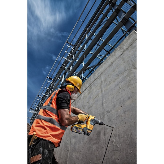 Worker in safety gear uses DEWALT drill on concrete wall at construction site under a clear sky.