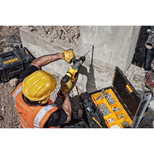 Worker with Dewalt drill making hole in concrete wall near toolboxes and fastener kit on construction site.
