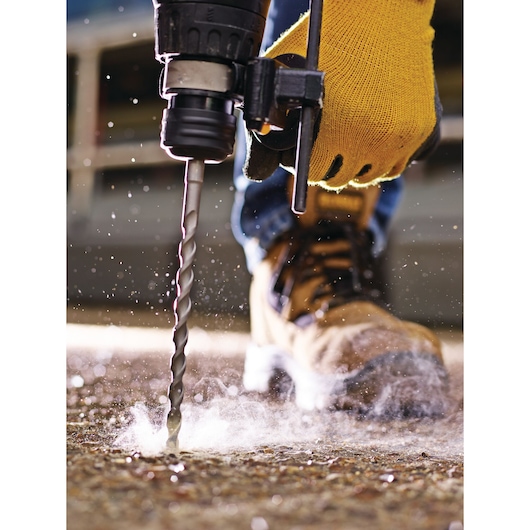 Gloved hand uses a DEWALT drill bit to drill into concrete, with dust and debris visible on the ground.