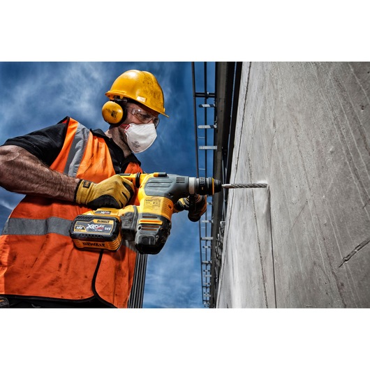 Worker uses a DEWALT cordless drill to bore into concrete wall outdoors, wearing safety gear and orange vest.
