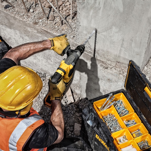 Worker drills into concrete using DEWALT DT9580, DT9580-QZ bits. Tool case with assorted fasteners is nearby.