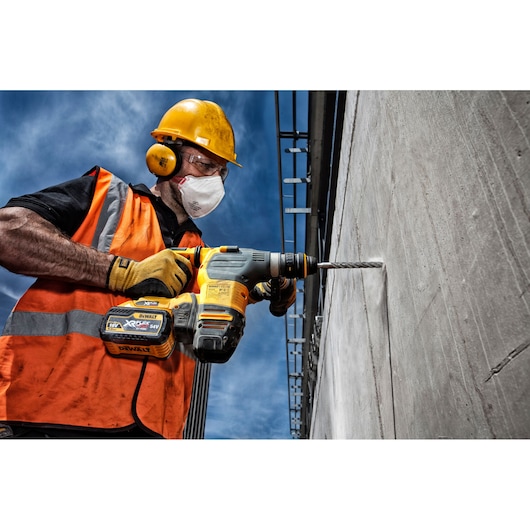 Worker uses DEWALT hammer drill to drill into a concrete wall outdoors, wearing safety gear and orange vest.