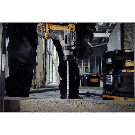 A DEWALT rotary hammer drill being used to drill into concrete at a construction site with storage boxes in the background.