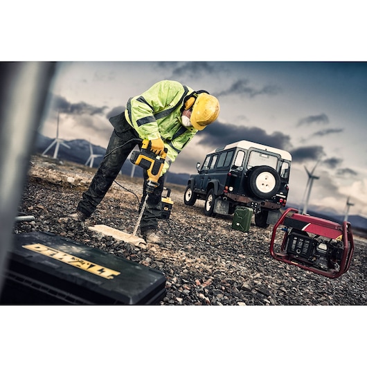 Worker drills into ground with DEWALT tool. Off-road vehicle and generator are nearby, wind turbines in background.