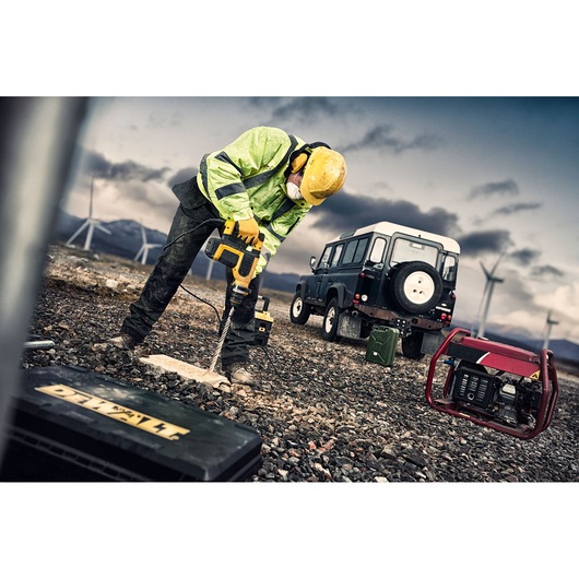 Worker drills stone outdoors with DEWALT tool, wearing high-vis gear and helmet. Vehicle and generator in background.