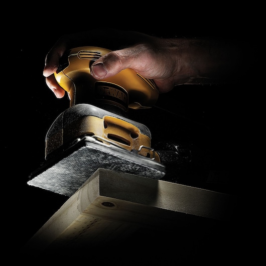 Hand holding a DEWALT sander, smoothing a wooden surface with the tool under focused lighting.
