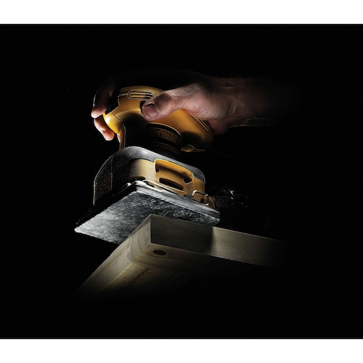 Hand using a DEWALT sander to smooth the edge of a wooden plank against a dark background.