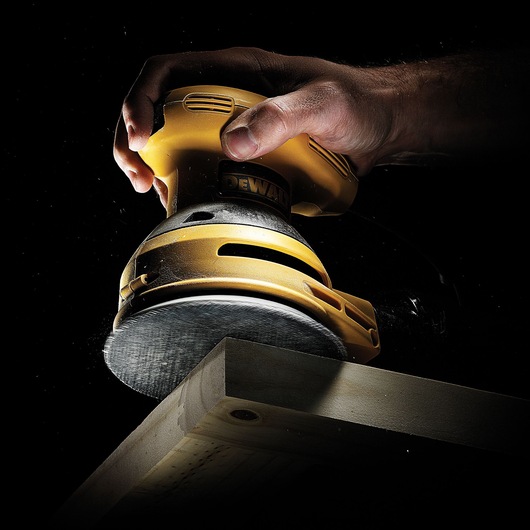 Hand holds DEWALT sander polishing wooden edge against dark background.