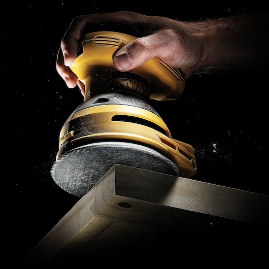 Hand holding a DEWALT sander, smoothing the edge of a wooden plank against a dark background.