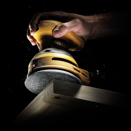 A DEWALT hand sander sanding a wooden plank, held firmly in a hand against a dark background.