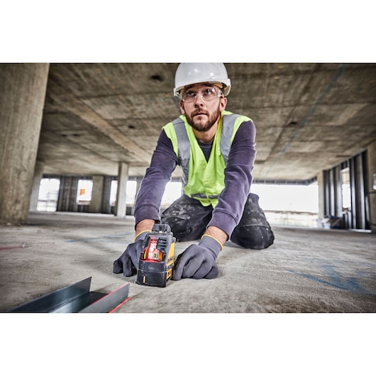 Worker using a DEWALT DW088K laser level on a construction site floor, wearing gloves and safety gear.