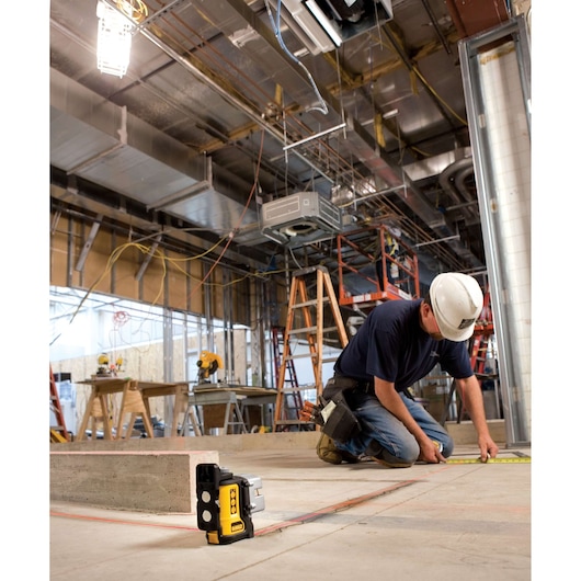 A DEWALT DW089K laser level is positioned on the ground at a construction site while a worker wearing a hard hat and tool belt measures with a tape measure on the floor. The background shows various construction equipment, ladders, and exposed ceiling pipes.