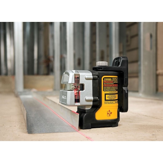 DEWALT DW089K three-beam laser level placed on a construction site floor, projecting a red laser line along a metal framing track with metal wall studs in the background.