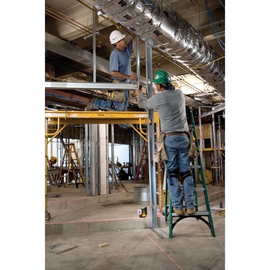 Two construction workers are assembling metal framing in an indoor construction site. One worker stands on a green ladder, while the other kneels on yellow scaffolding. A DEWALT DW089K laser level is placed on the floor below, projecting red laser lines to assist with alignment. The workspace is filled with construction equipment, ductwork, and scaffolding.