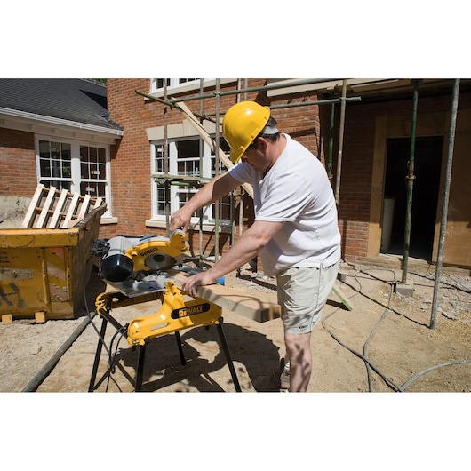 Person using a DEWALT DW743N saw to cut wood at a construction site with red brick buildings in the background.