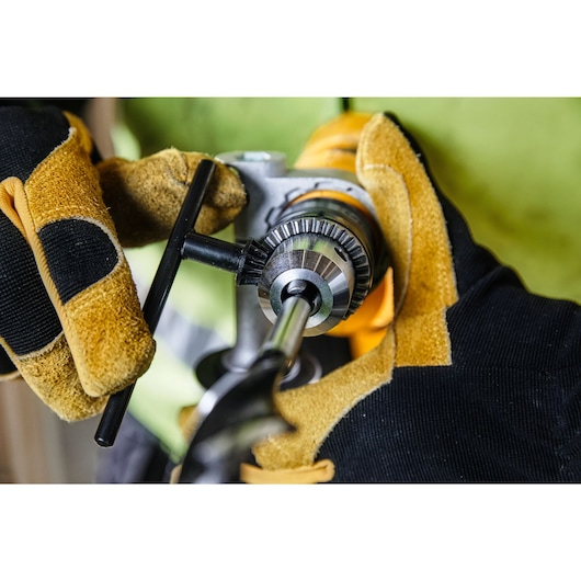 Gloved hands adjust a DEWALT drill with a chuck key for a drill bit. Close-up of tool and safety gloves.