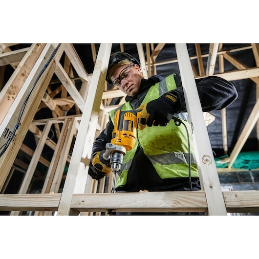 DEWALT DWD024K drill in use on wooden beams at a construction site by a person wearing safety gear.