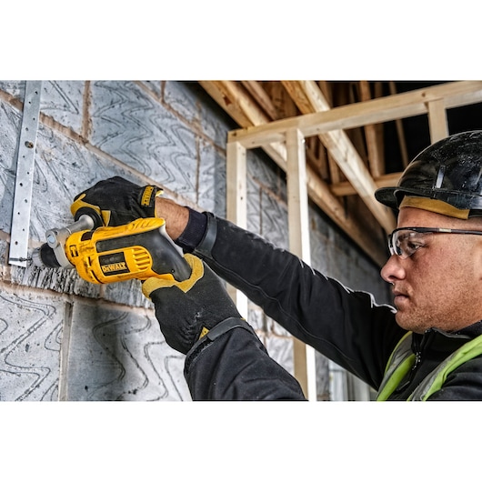 Gloved worker uses DEWALT drill DWD024K on a brick wall at a construction site. Face is blurred for privacy.